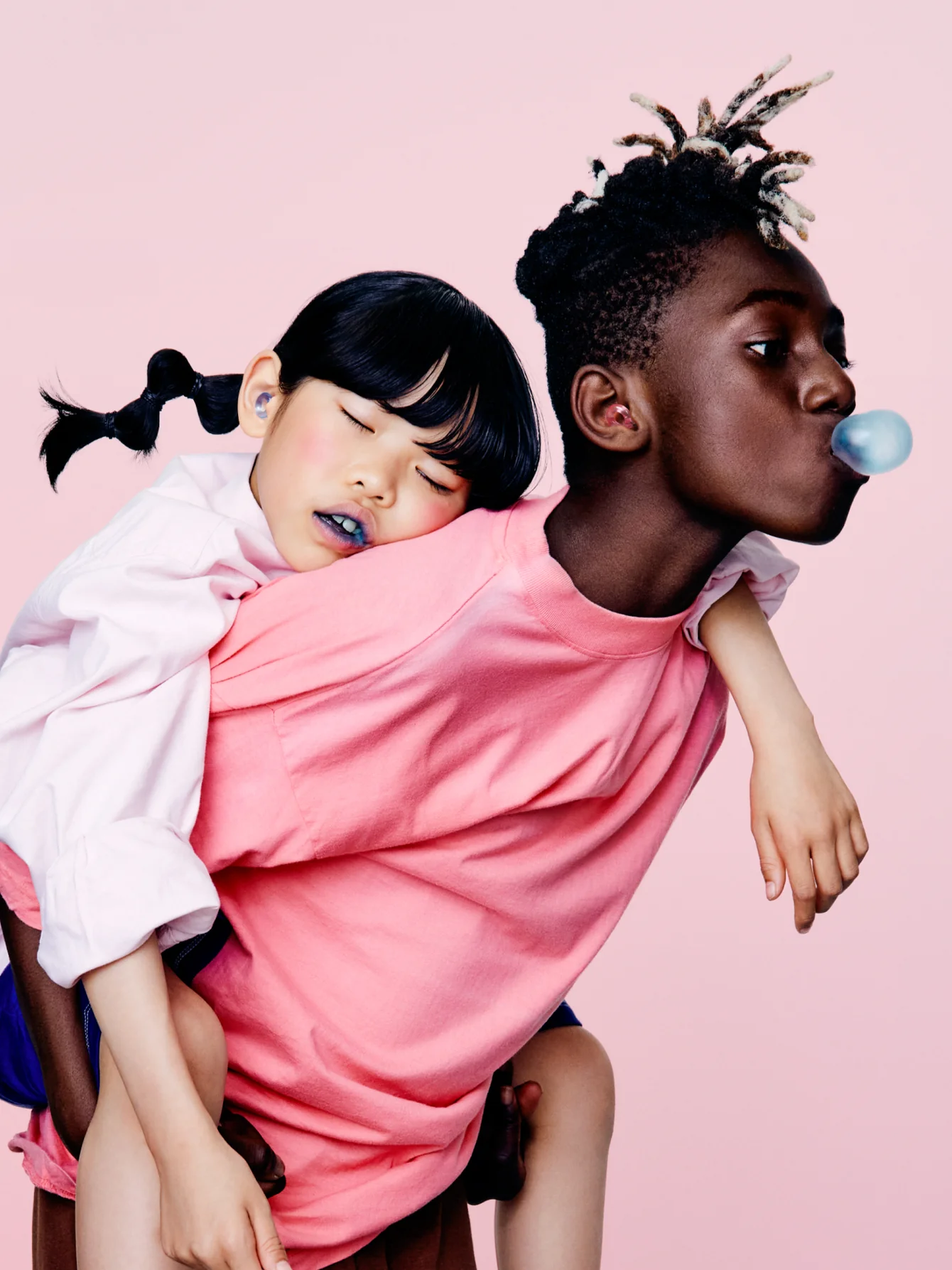 A young boy with short, twisted hair and a pink shirt gives a piggyback ride to a girl with black pigtails, who is sleeping with blue lipstick on. He blows a blue bubblegum bubble against a pink background.